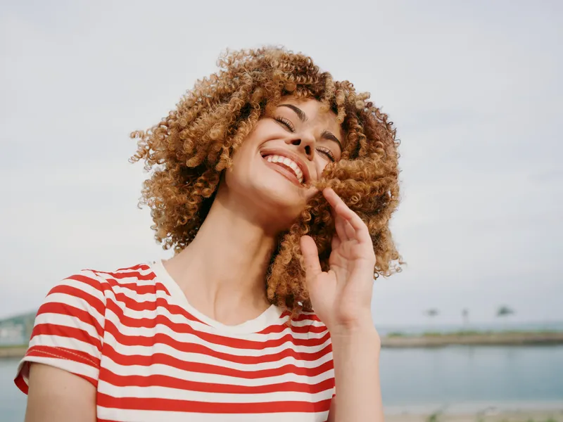 Joyful portrait of a young woman with curly hair wearing a red and white striped shirt, smiling brightly against a soft outdoor backdrop, radiating warmth and confidence.