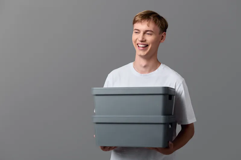 Smiling boy holding a stack of gray storage boxes in a casual white tee, clean studio background, demonstrating tidy storage, packaging and organization for home or workspace