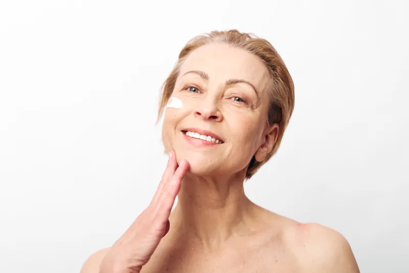 Mature smiling woman applies white facial cream on clean skin against white background. Concept of skincare, antiaging, selfcare, beauty routine, and natural glow for healthy skin.