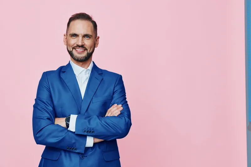 A confident business professional stands with arms crossed against a solid pink backdrop, wearing a blue suit and white shirt. The pose conveys poise, professionalism, and approachability.