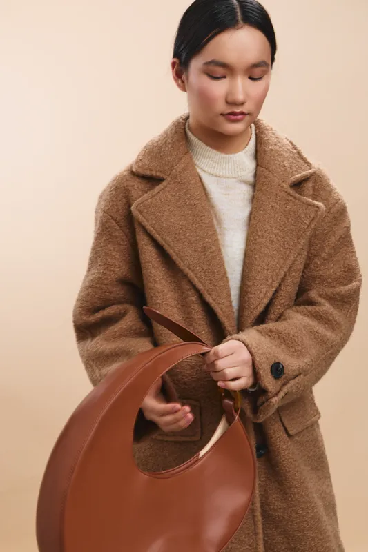 asian woman wearing a warm brown coat stands against a soft neutral backdrop, gazing downward at a leather handbag with calm, graceful poise and modern fashion appeal.
