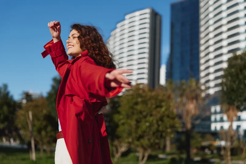 Cheerful woman outdoors, arms outstretched, wearing a red coat, sunny day, carefree pose, vibrant colors, lush greenery with urban skyline backdrop
