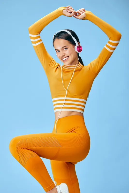 Fitness woman in bright orange activewear stretches arms overhead in a studio, listening to headphones for a vibrant, energetic workout and dance-inspired routine.