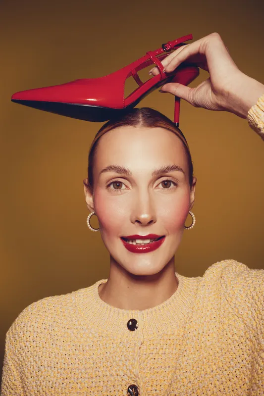 Young woman with red lipstick and hoop earrings holds red high heel shoe above her head against brown background. Fashion concept with stylish makeup and confident expression.