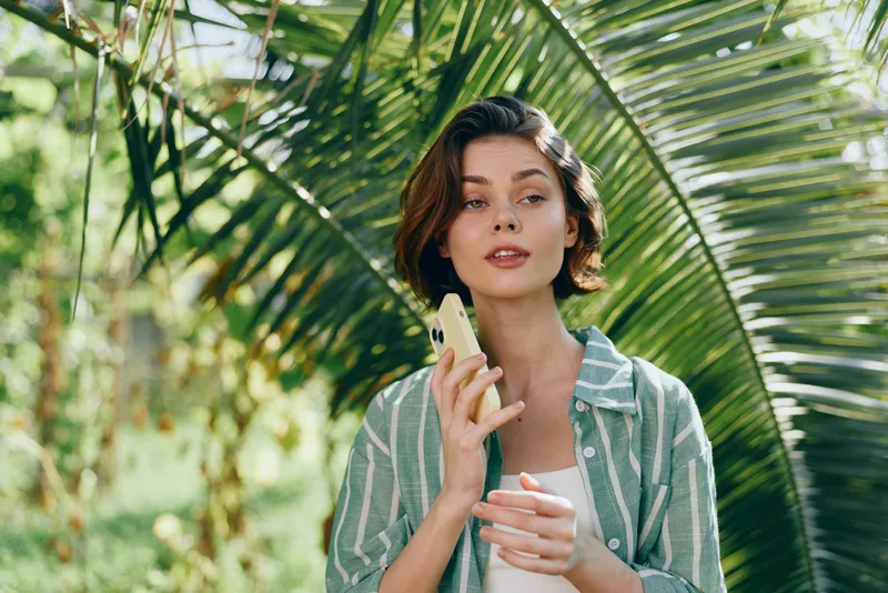 Main focus on a stylish young woman outdoors holding a smartphone amid tropical greenery, with soft sunlight and a relaxed expression.