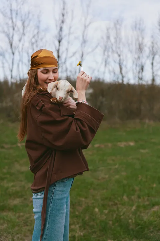 Cheerful woman outdoors, smiling with a small dog, sunlit moment, relaxed posture, soft breeze, natural colors, grassy field, bare trees in the background, candid lifestyle scene