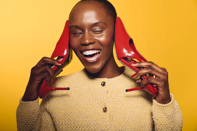 Happy dark skinned African woman wearing beige sweater holds red high heels near ears, smiling with closed eyes on yellow background. Concept of joy, fashion, and beauty with colorful contrast.