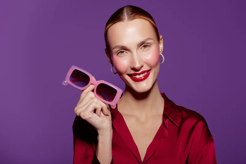Smiling woman in red blouse holds pink sunglasses near face against purple background. Studio portrait with bright makeup and hoop earrings. Concept of fashion, style, and beauty accessories.