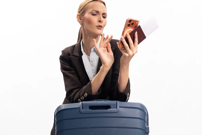 A serious woman in a dark jacket holds a smartphone and passport while standing behind a blue suitcase. The image illustrates travel, planning, and preparation concepts.