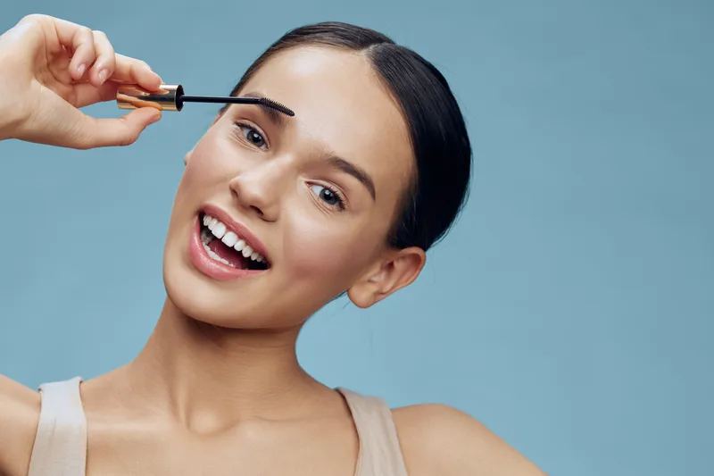 Young woman applying eyebrow gel, showcasing her beauty routine with a joyful expression against a pastel blue background, highlighting confidence and self care