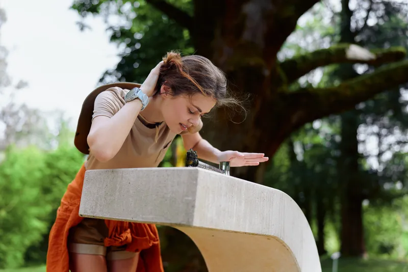 Girl drinking water from a public fountain outdoors in park, leaning over concrete basin with backpack and casual summer clothing, refreshing moment under trees and sunlight.
