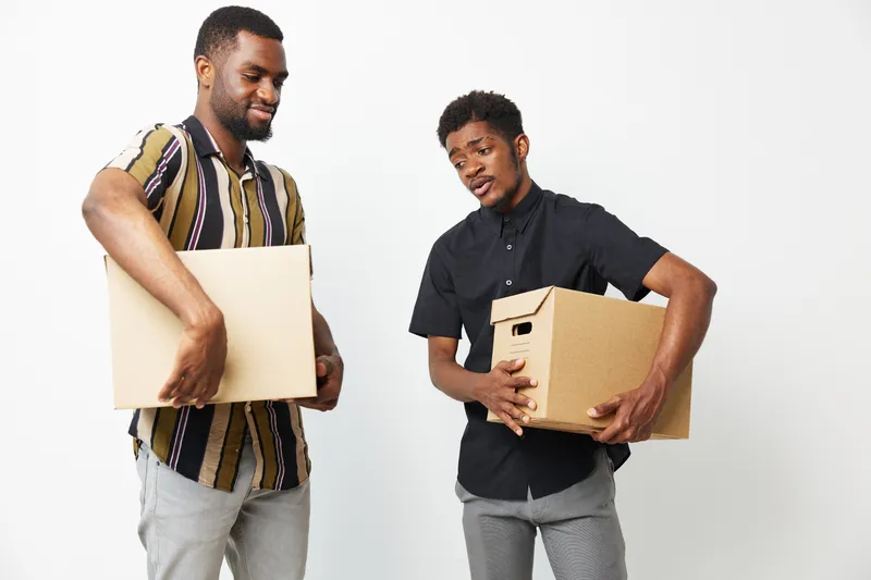 Two African American men carrying boxes, showcasing teamwork and collaboration while smiling. A bright and dynamic image representing partnership and positivity.