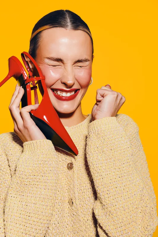 Happy woman in yellow sweater holds red high heel shoe near face, eyes closed and smiling. Bright orange background creates vibrant and joyful mood, concept of fashion and excitement.