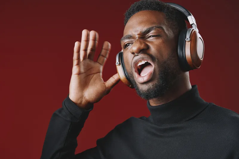 Young Black man enjoying music with headphones, expressing joy and passion against a vibrant red background wearing a black turtleneck