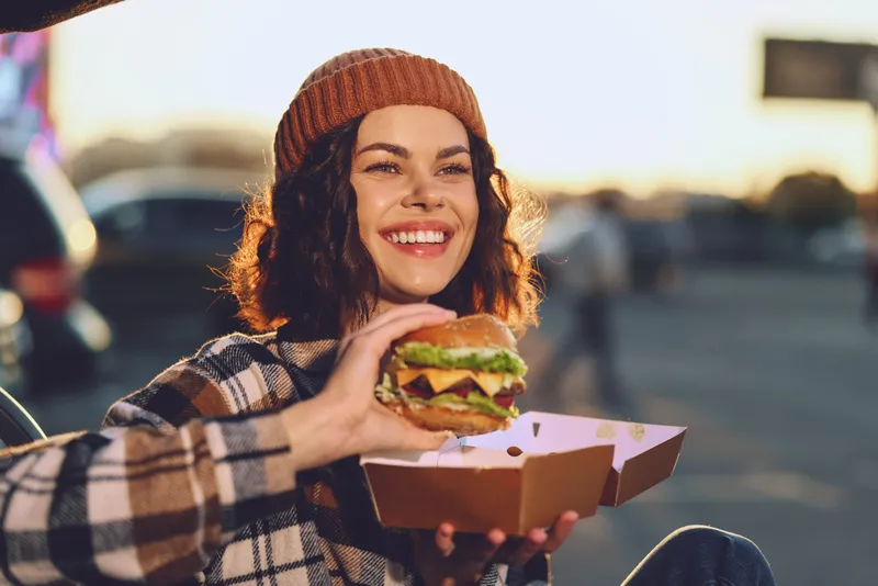 woman holding burger with bright smile outdoors at golden hour glow, candid authenticity and mindful living mood, emotional storytelling of urban food joy and relaxed lifestyle