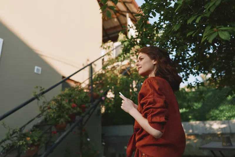 Cheerful woman outdoors, side profile, warm sunlight, rust dress, relaxed posture, soft breeze, lush greenery, natural colors, lifestyle moment, candid atmosphere