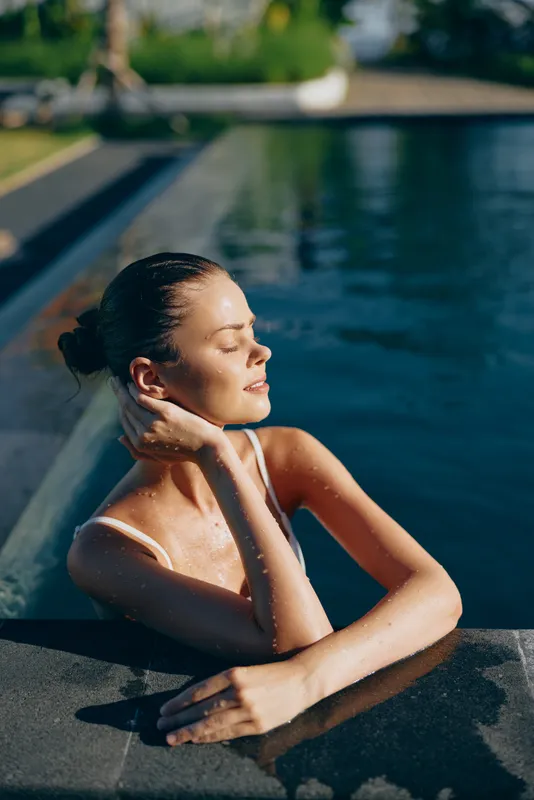 A woman rests at the pool edge, eyes closed, enjoying warm sunlight. The calm water and outdoor scene convey relaxation, leisure, and a moment of peaceful solitude.
