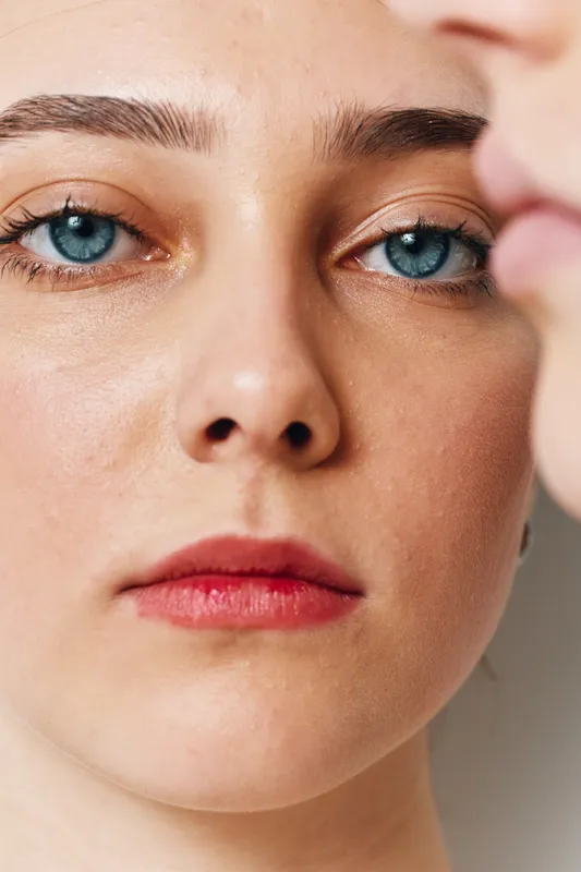 Closeup of woman face with blue eyes and natural skin texture in minimal makeup showing emotion and beauty with soft light and blurred profile in background.