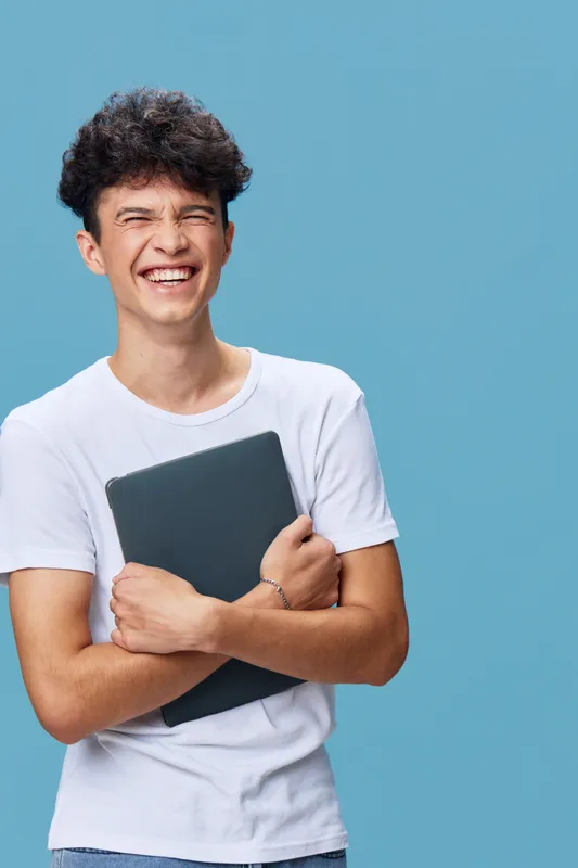smiling man holding laptop stands against blue background, wearing casual white t-shirt, cheerful young adult with curly hair, bright and positive expression