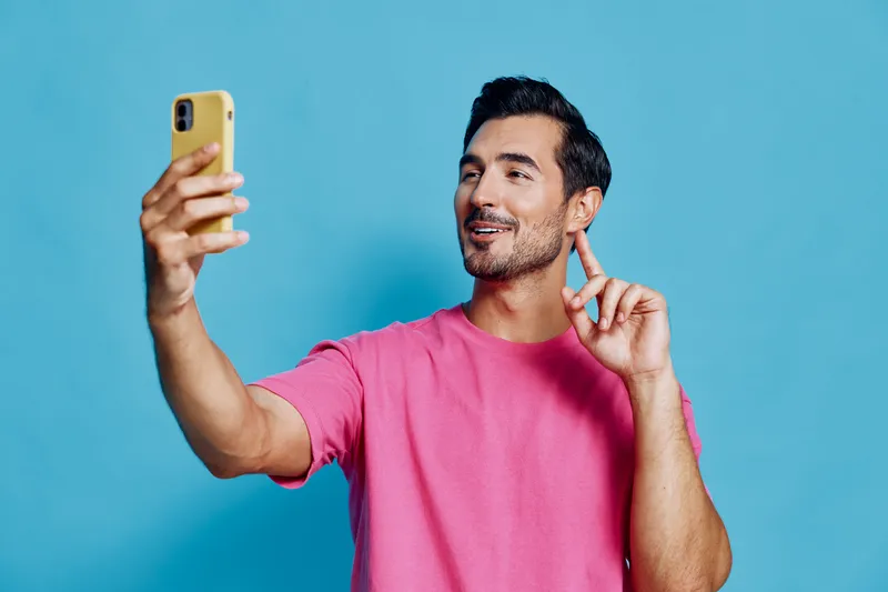 Cheerful smiling man taking a selfie with his mobile phone, wearing a bright pink shirt, against a solid blue background, capturing a joyful moment in his life.