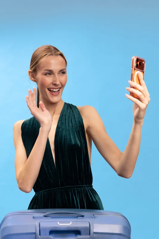 Young woman in elegant dress waves and smiles during video call while standing behind suitcase on blue background. Concept for travel, communication, and remote connection.