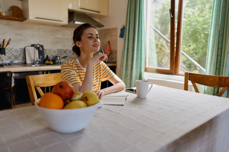 A thoughtful woman sits at a kitchen table, hand on chin, gazing out the window. Warm light fills the room, creating a calm moment of reflection, routine, and personal care.
