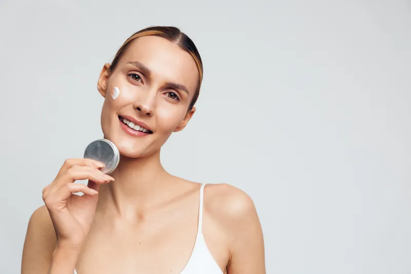 Smiling woman applying white face cream on cheek with a small container in hand. Close-up on a white background. Concept of skincare, beauty, and self-care routine with natural light.