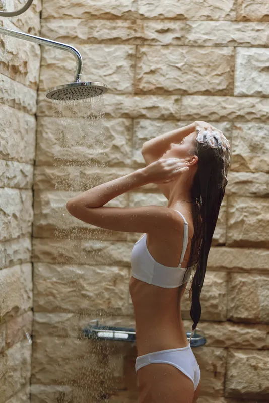 young woman washing hair under outdoor shower with stone wall background, natural light, summer wellness and skincare routine concept