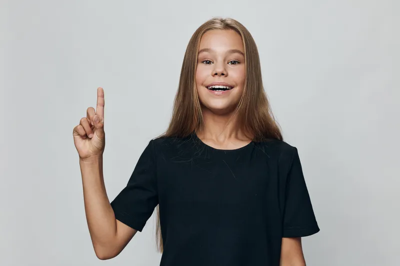Girl with long hair wearing black t-shirt smiling and pointing up with forefinger against neutral background in studio. Youthful child showing gesture and happy expression.