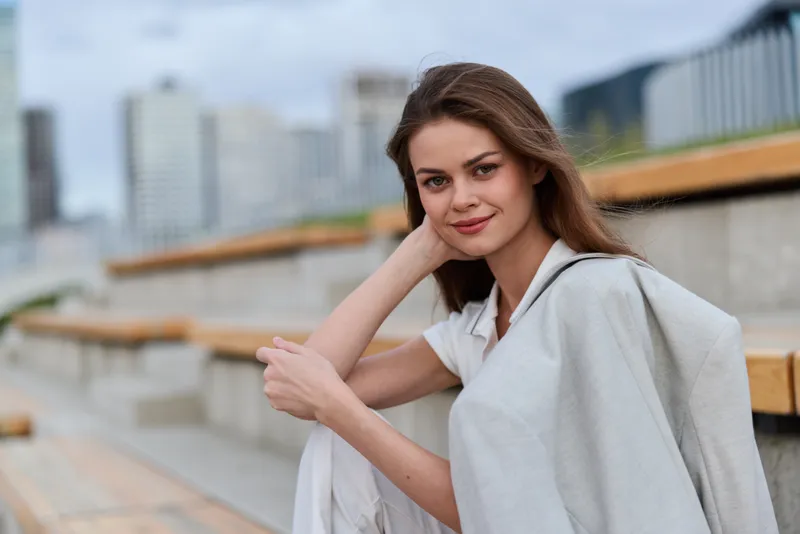 Young woman with light brown hair in casual gray outfit sitting outdoors on urban wooden benches with city skyline background, calm and confident expression