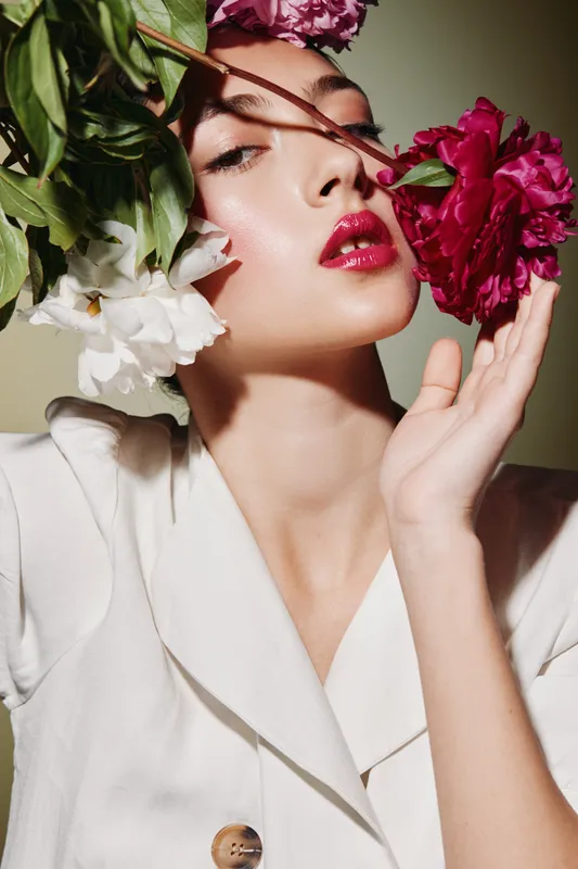 Fashion model with glossy red lips embraces vibrant pink and white flowers near her face. Creative beauty shoot focusing on natural elegance and fresh skin in soft lighting.