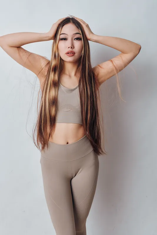 Young woman with long straight hair in fitness outfit posing with hands on head against white wall for wellness and beauty concept.