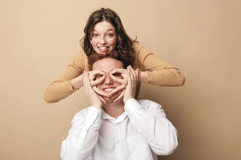 Happy young couple having fun together, woman playfully covering man's eyes with her hands. Studio portrait on beige background, concept of love, joy, friendship, and positive emotions.