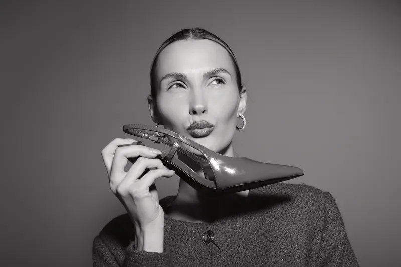 White woman with light skin holds shiny high heel shoe near face, making playful kiss gesture in studio. Concept of fashion, style, and beauty accessories.