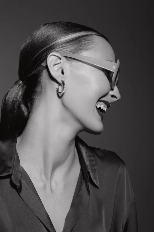 Smiling Caucasian woman with pale skin and sunglasses laughs with head turned sideways in black and white studio portrait.