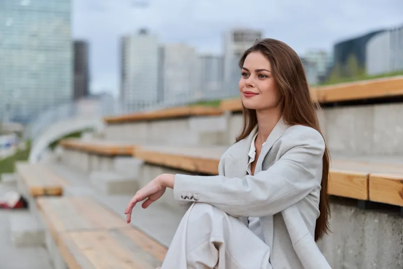 confident woman in light business suit sitting on urban wooden bleachers with relaxed smile, long brown hair, outdoor city background, daytime, fashion style