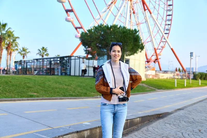 Woman outdoor wheel fashion casual jeans jacket smile street park in an urban setting beside a ferris wheel, sunny day, travel style and street fashion in a relaxed park environment