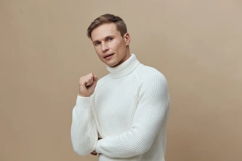 Confident young man wearing cozy white turtleneck sweater looking directly at camera posing with fist slightly raised isolated on beige background studio portrait. People lifestyle concept