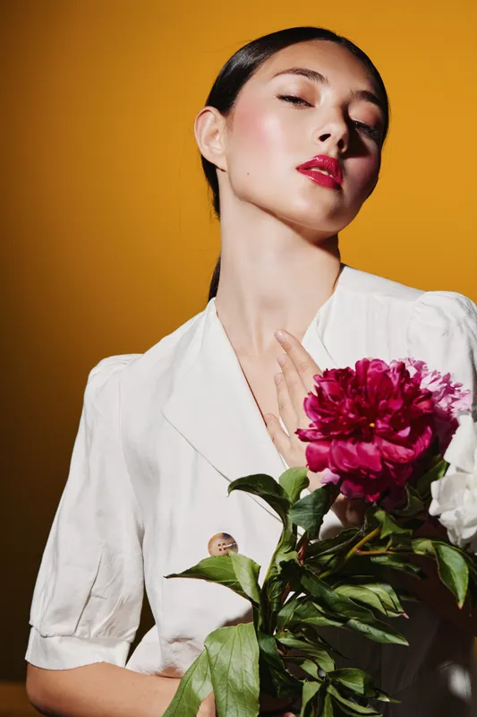 Fashion model with sleek hairstyle and glossy red lips poses elegantly with deep pink peony flower against a vibrant yellow background. Beauty, creative, expressive mood.