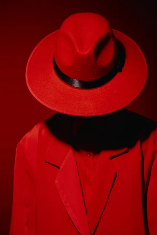 Person dressed in all red fashion attire with wide brim hat shadows face against red backdrop. Creative beauty concept focusing on bold color and dramatic style.