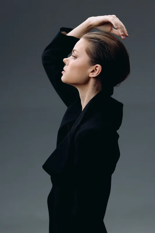 Profile of a calm woman in black clothing with raised arm, elegant pose, and neutral gray background.