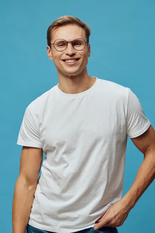 Smiling man wearing glasses and white t-shirt posing on blue background. Studio portrait demonstrating happiness, positivity, casual style, cheerful mood, young adult male, friendly expression, relaxe