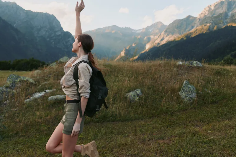 A woman with a backpack and baby carrier stands in a mountain meadow, reaching up with one hand. The scene captures outdoor lifestyle and nature exploration.