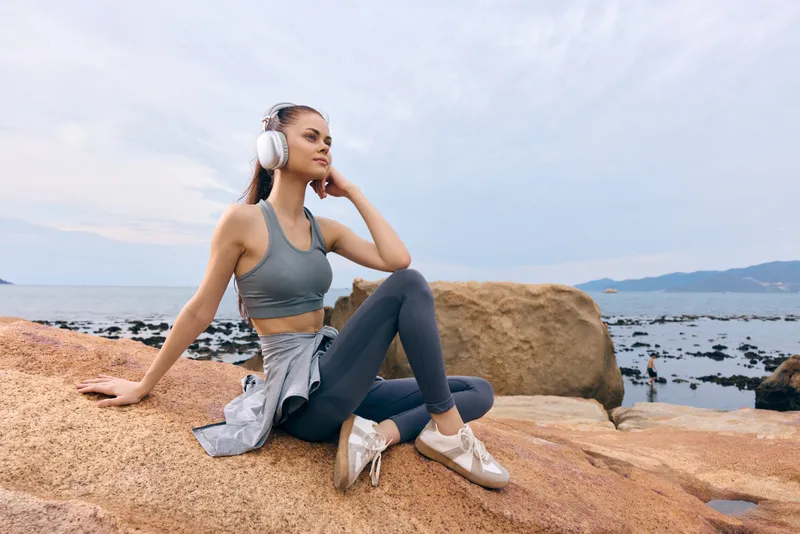 Young woman enjoying music outdoors, wearing activewear, sitting on rocks by the sea, calm expression, healthy lifestyle, relaxation
