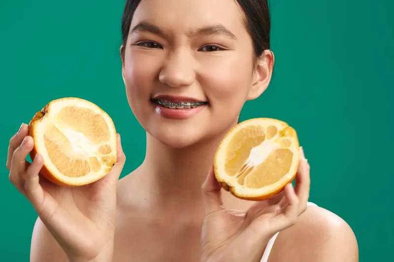 Asian woman holds lemon halves against a vivid green background, smiling warmly with braces showing. Bright, fresh mood and citrus vibe convey health and vitality.