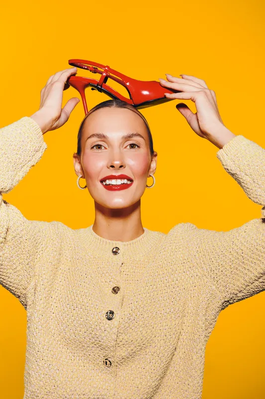 Smiling woman in beige sweater holds red high heel shoe above head against bright yellow background. Concept of fashion, style, happiness, and colorful modern portrait.