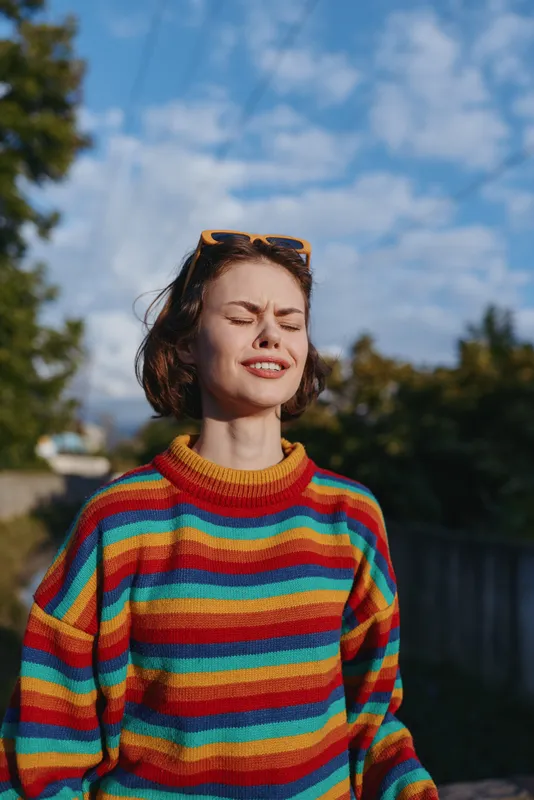 Young woman portrait smiling with eyes closed in rainbow sweater outdoors, short brown hair and sunglasses on head, relaxed sunny suburban scene for lifestyle travel and joy.