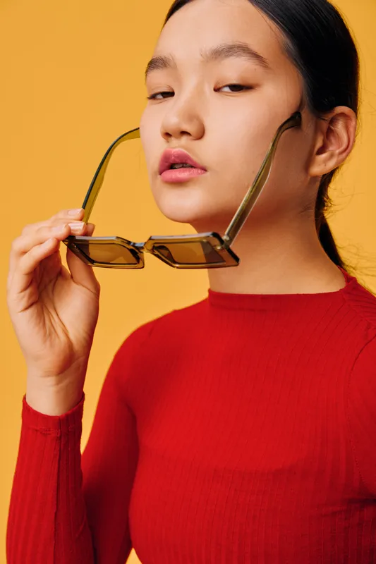 Young woman holding sunglasses near her face with serious expression wearing red long sleeve shirt on yellow background, portrait with fashion and style concept for modern look.