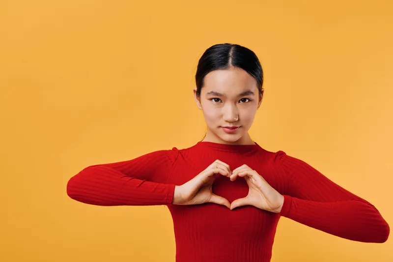 Young woman in red sweater making heart shape with hands against yellow background. Portrait of female showing love and care gesture with confident expression and neat hairstyle.