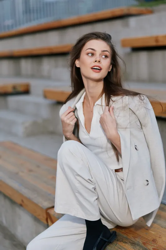 Stylish businesswoman exuding confidence, dressed in a tailored light suit, sitting on wooden steps with a relaxed smile and a sophisticated look.